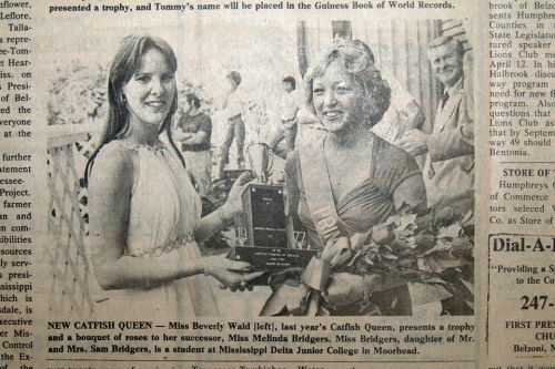 Two women in formal attire smile while holding a trophy and bouquet, celebrating a Catfish Queen title.