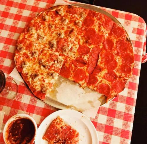 A large pizza with pepperoni and sausage, served on a checkered tablecloth, with a slice and marinara sauce on the side.