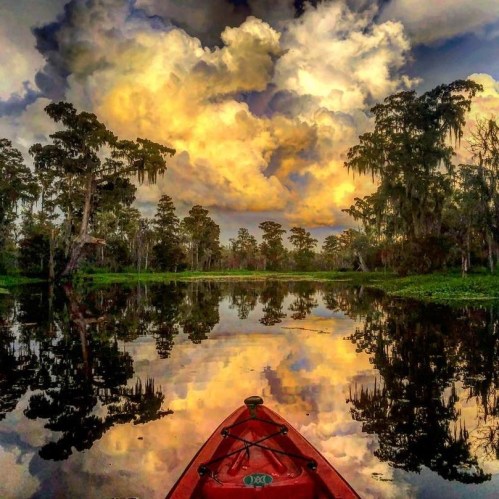 A kayak on a calm lake reflects vibrant clouds and lush trees under a colorful sky at sunset.