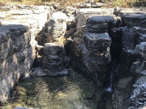 Rocky formations surround a clear pool of water, with sunlight filtering through the trees above.