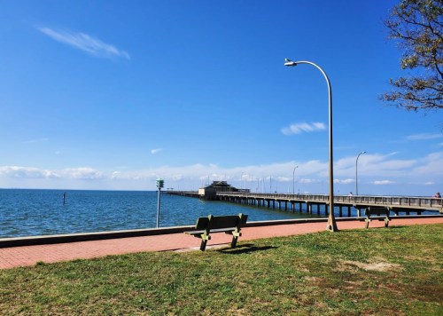 A serene waterfront view featuring a pier, benches, and clear blue skies over calm water.
