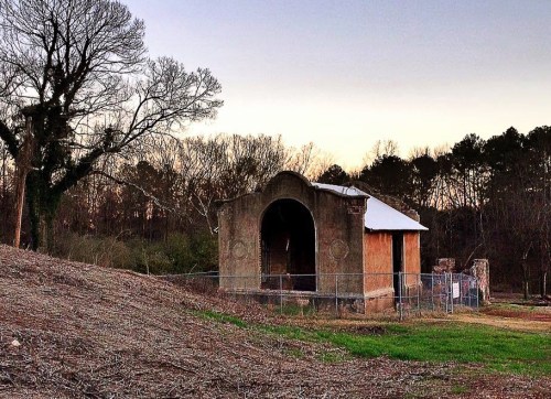 An old, abandoned structure with an arched entrance, surrounded by trees and a fenced area at dusk.
