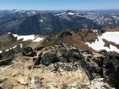 A rocky mountain peak with snow patches, overlooking a vast landscape of rugged mountains and clear blue skies.