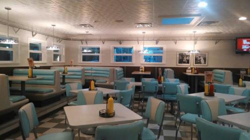 A retro diner interior with teal booths, checkered floor, and tables, featuring condiments on each table and framed photos on the walls.