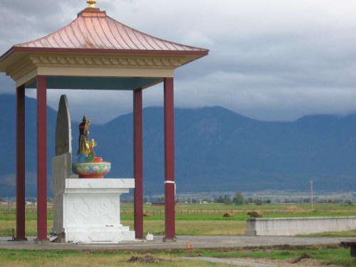 A colorful statue of a deity under a pavilion, set against a backdrop of mountains and cloudy skies.