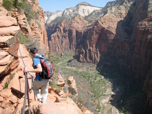 A hiker navigates a rocky trail along a steep canyon, with stunning red rock formations and a river below.