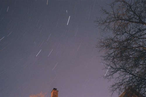 A night sky with star trails, featuring a silhouette of a tree and a chimney in the foreground.