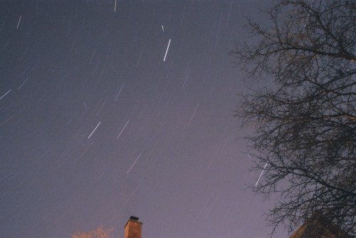 Star trails streak across a night sky, with a silhouette of a tree and a chimney in the foreground.