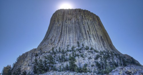 A towering rock formation with vertical stripes, topped by a bright sun against a clear blue sky.