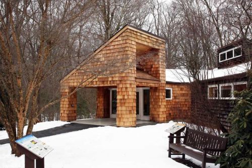 A modern wooden building with a unique design, surrounded by snow and bare trees. Benches and informational signs are nearby.