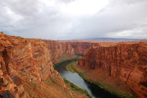 A winding river flows through a dramatic canyon with steep, reddish cliffs under a cloudy sky.