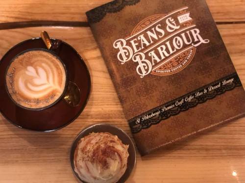 A cup of latte with latte art beside a cinnamon roll, next to a menu for "Beans & BarLOUR" on a wooden table.