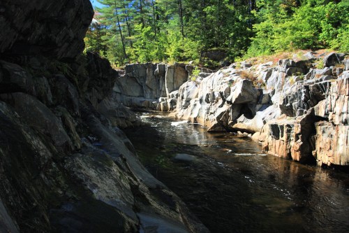 A serene river flows between rocky cliffs, surrounded by lush green trees under a clear blue sky.