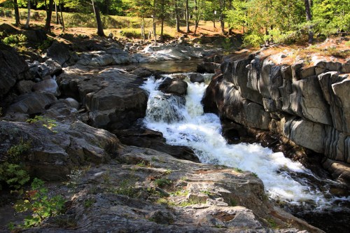 A serene waterfall cascades over rocky terrain, surrounded by lush greenery and trees in a peaceful natural setting.