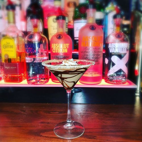 A chocolate-drizzled cocktail in a martini glass, with colorful vodka bottles in the background.