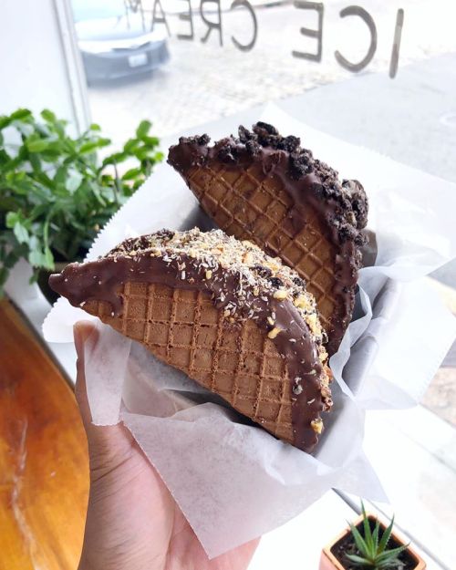 A hand holding two chocolate-dipped waffle cones filled with ice cream and topped with crushed cookies and nuts.