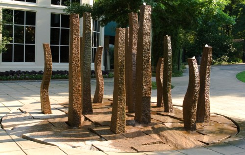 A modern sculpture featuring tall, textured metal pillars arranged in a circular formation on a stone patio.