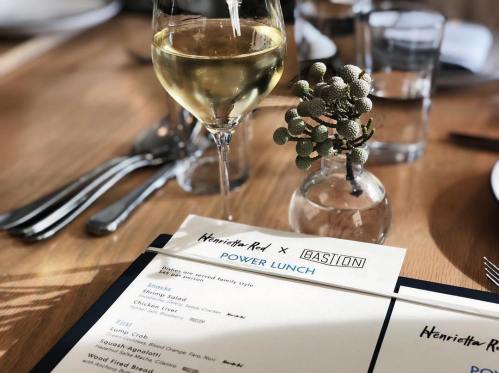 A glass of white wine beside a small vase and a menu for a power lunch at Henrietta Red x Bastion.