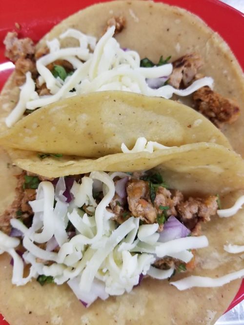 Two tacos filled with seasoned meat, chopped onions, cilantro, and topped with shredded cheese on a red plate.