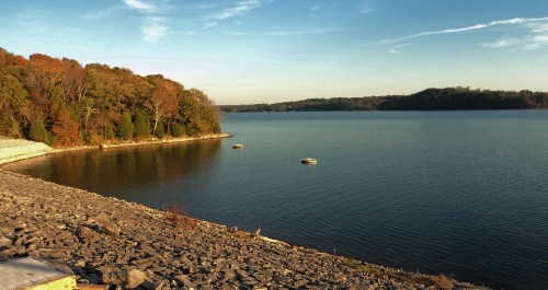 A serene lake scene with calm water, rocky shore, and trees displaying autumn colors under a clear blue sky.