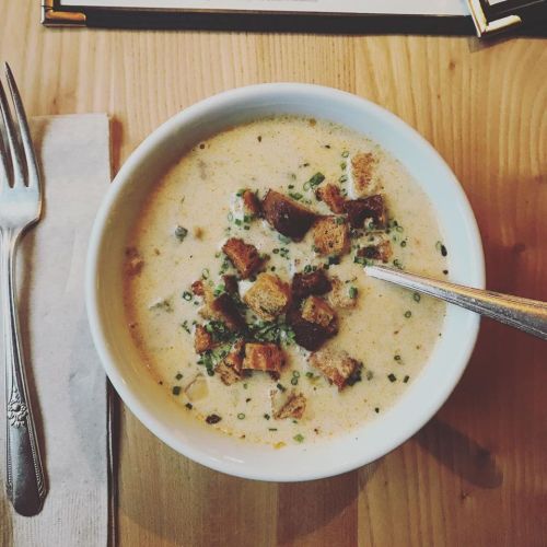 A bowl of creamy soup topped with croutons and herbs, placed on a wooden table with a fork and napkin nearby.