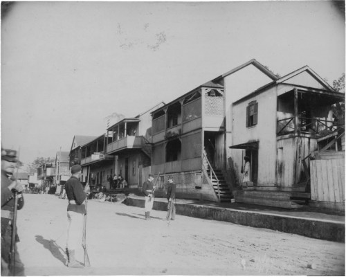 Historic black and white photo of damaged buildings, with soldiers and civilians in a street scene.