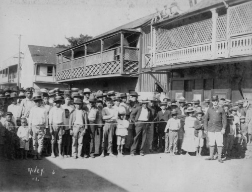 A large group of people, including children and adults, gathered outdoors in front of buildings, some wearing hats.