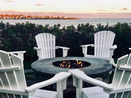 A cozy outdoor seating area with white chairs around a fire pit, overlooking a calm waterfront at sunset.