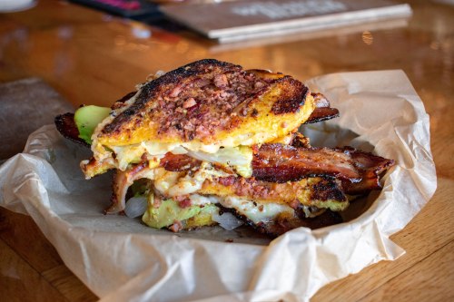 A stacked sandwich with crispy edges, filled with layers of meat, cheese, and pickles, served in a paper-lined basket.