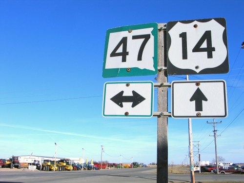 Road signs for highways 47 and 14, with directional arrows, against a clear blue sky and machinery in the background.