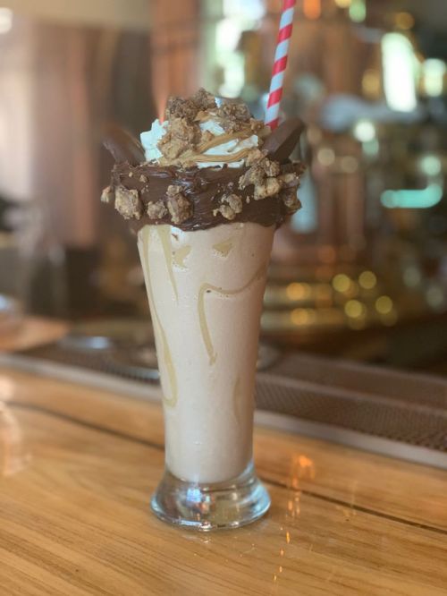 A tall milkshake topped with whipped cream, chocolate, and crunchy toppings, served in a glass with a striped straw.