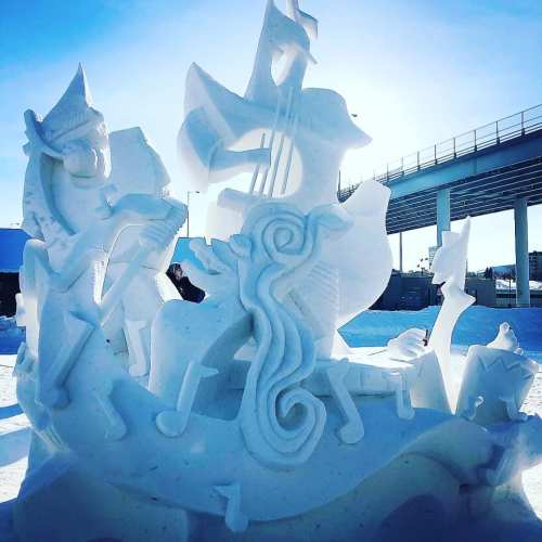 A detailed snow sculpture featuring musicians and animals, surrounded by musical notes, under a bright blue sky.