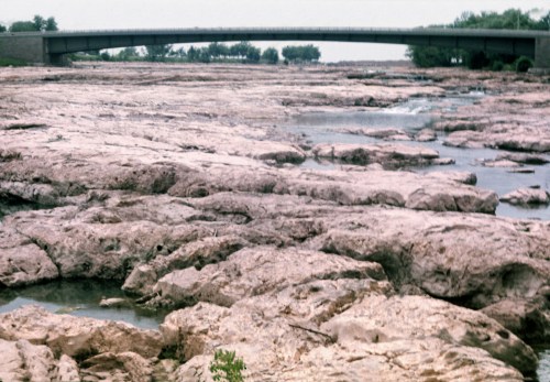A rocky riverbed with sparse water and a bridge in the background, surrounded by trees under a cloudy sky.