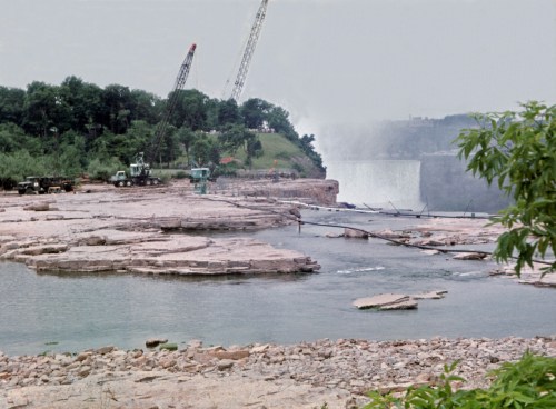 Construction equipment near a riverbank, with cranes and a waterfall in the background, surrounded by greenery.