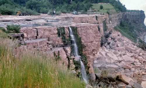 A rocky cliff with a waterfall, surrounded by greenery and machinery, showcasing a rugged landscape.