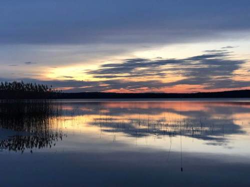A serene lake at sunset, reflecting colorful clouds and silhouettes of reeds along the shore.