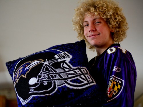 A smiling person with curly hair holds a decorative pillow featuring a football helmet design.