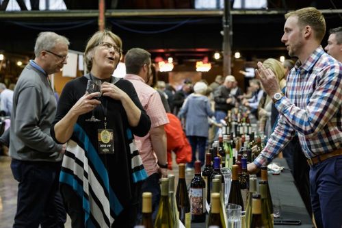 A woman talks animatedly while holding a glass of wine at a busy wine tasting event with various bottles on display.