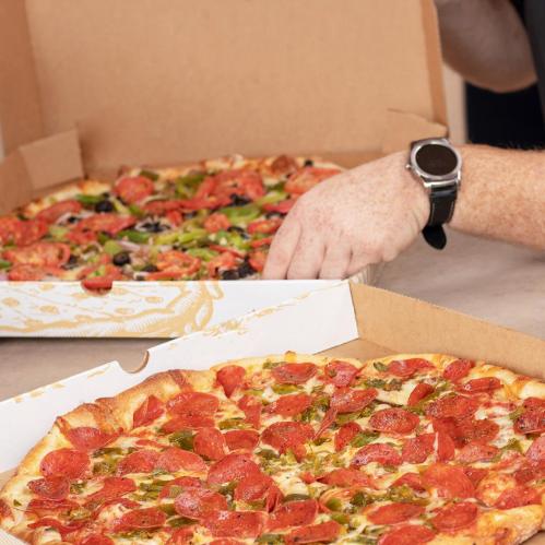 A hand reaches for a pepperoni pizza in a box, with another pizza featuring toppings in the background.
