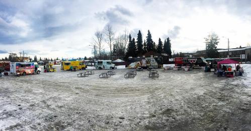 A snowy outdoor food truck gathering with various trucks and picnic tables set up in a circular arrangement.