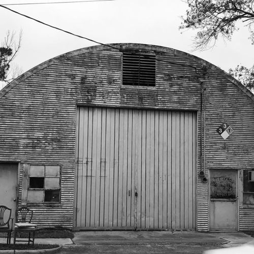 A weathered, corrugated metal building with a large door, small windows, and a chair in front, captured in black and white.