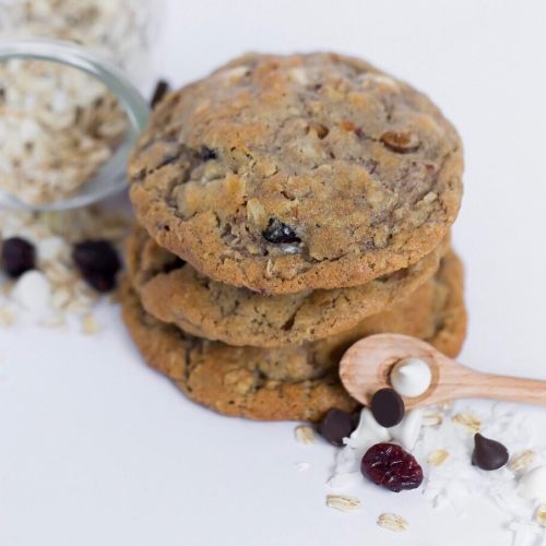 Three stacked cookies with chocolate chips and dried fruit, surrounded by oats and coconut flakes on a white background.