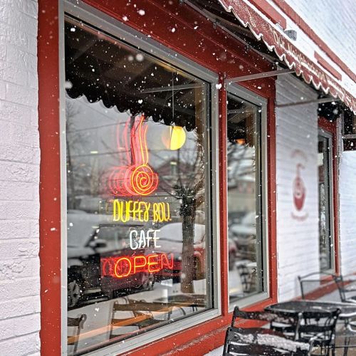 A cozy café window with a neon "OPEN" sign, surrounded by falling snow and outdoor seating.