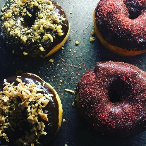 Four chocolate-glazed donuts topped with various sprinkles and nuts on a textured black surface.