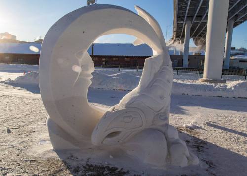 A snow sculpture featuring a stylized fish and a circular design, set against a snowy landscape and blue sky.