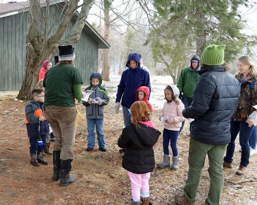 A group of adults and children in winter clothing gather outdoors, listening to a speaker near a cabin.