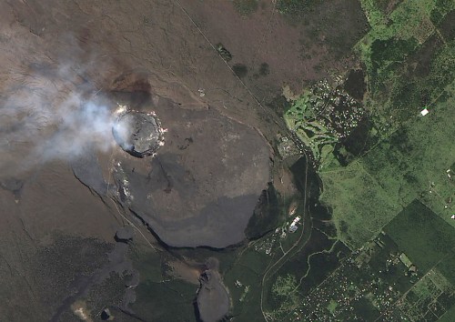 Aerial view of a volcanic crater emitting smoke, surrounded by lush green land and a nearby village.