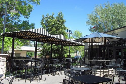 Outdoor dining area with striped canopies, black tables, and chairs surrounded by greenery under a clear blue sky.