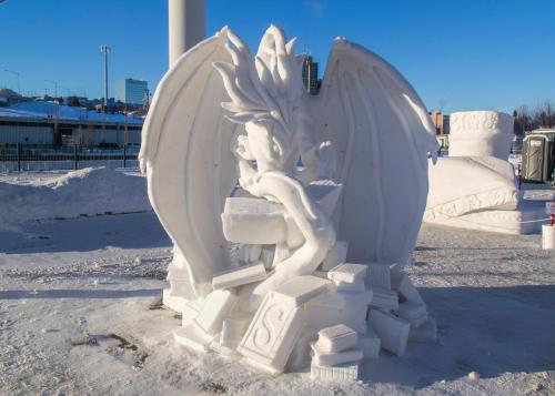 A detailed snow sculpture of a dragon-like creature surrounded by books, set against a clear blue sky.