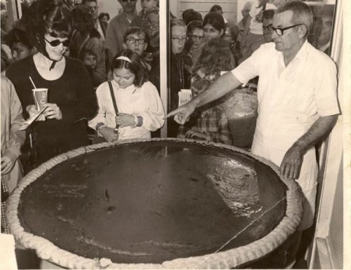 A man in a white apron serves a large dessert while a woman in sunglasses and a girl watch, surrounded by a crowd.